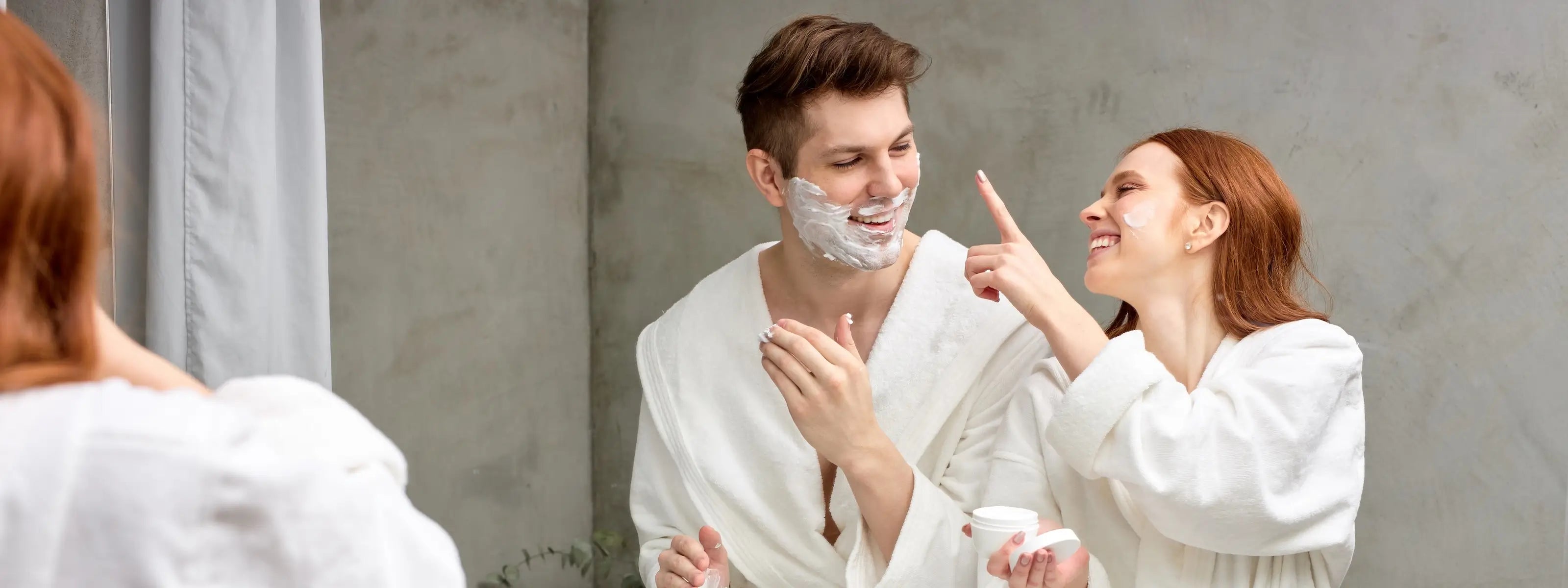 A man and a woman in white robes standing in front of a mirror, with the man shaving his face and the woman pointing at it. The man has shaving foam on his face.